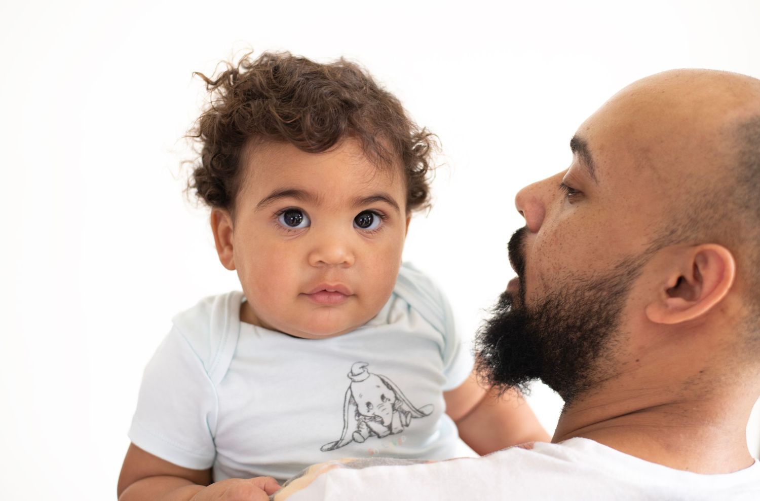 A bearded man in profile view holds a young child wearing a white t-shirt against a white background.