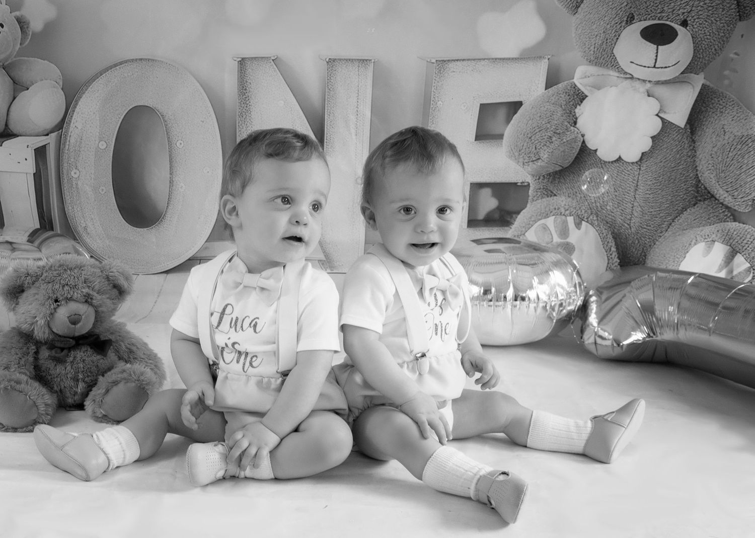 Black and white photo of twins sitting with LOVE letters and teddy bears in studio setting.