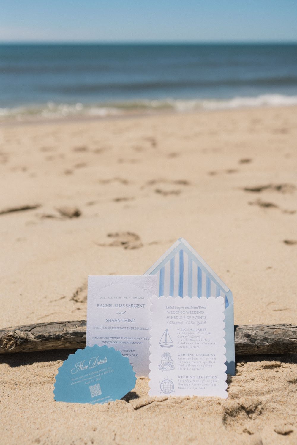Beach wedding invitations with nautical design, blue striped envelope liner, and scalloped edges displayed in sand by the ocean.