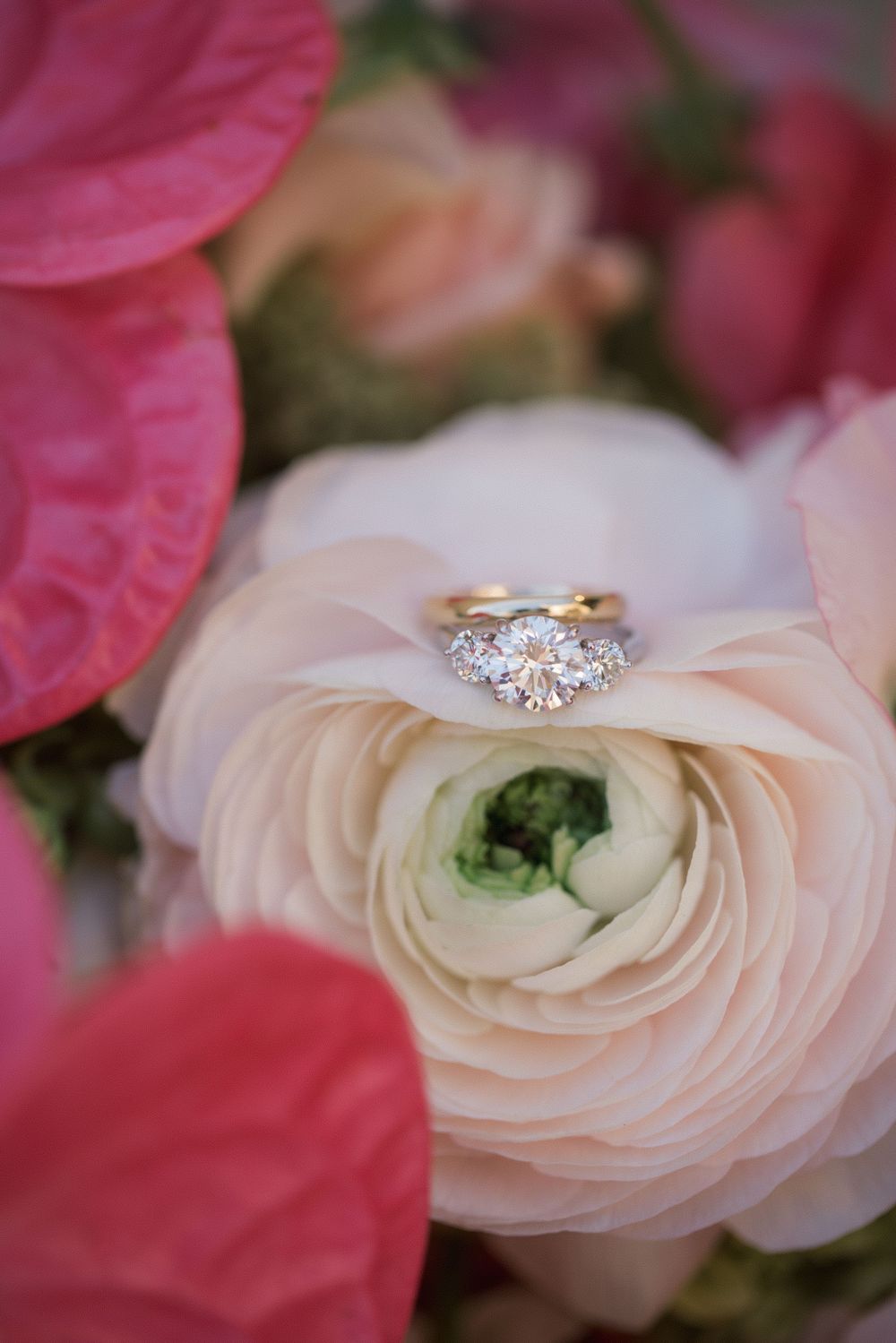 Diamond three-stone engagement ring resting on a blush pink ranunculus flower in a romantic floral bouquet.
