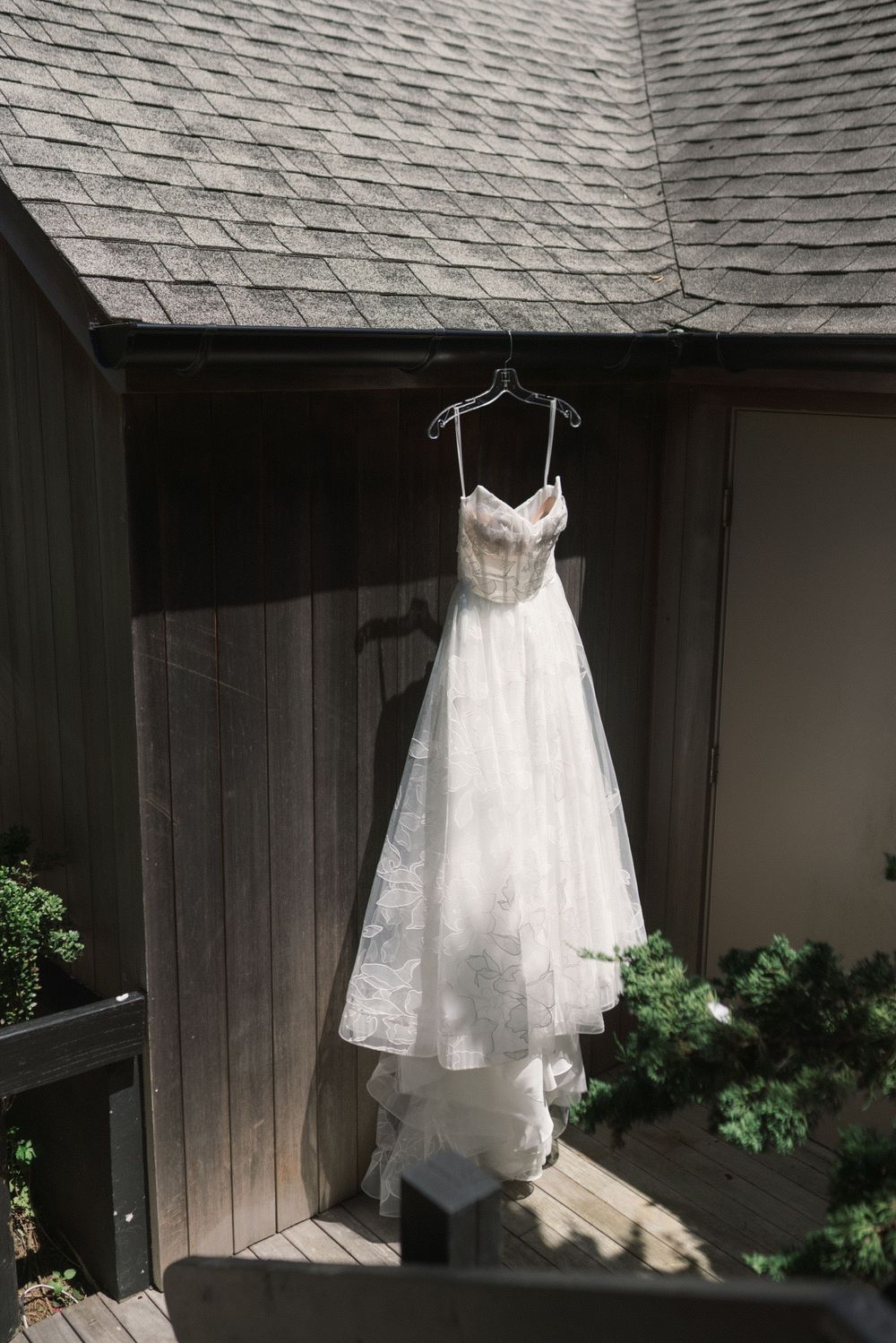 Elegant white lace wedding dress hanging on a hanger outside a rustic wooden building with a shingle roof.