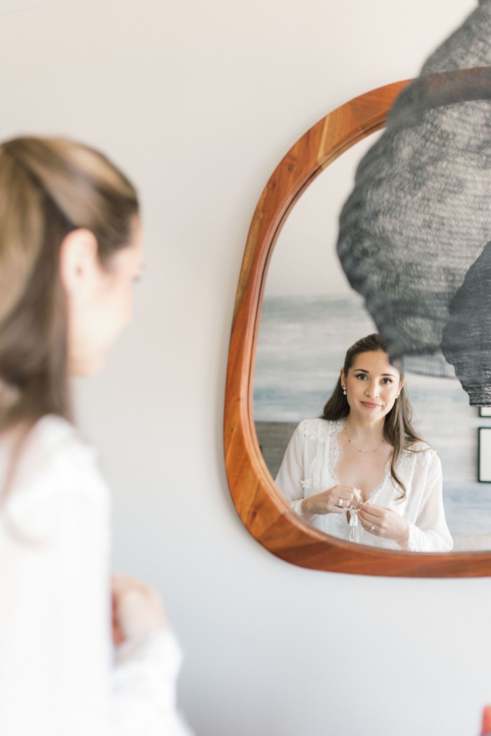 Bride in white lace robe looking at her reflection in a wooden framed mirror while getting ready.