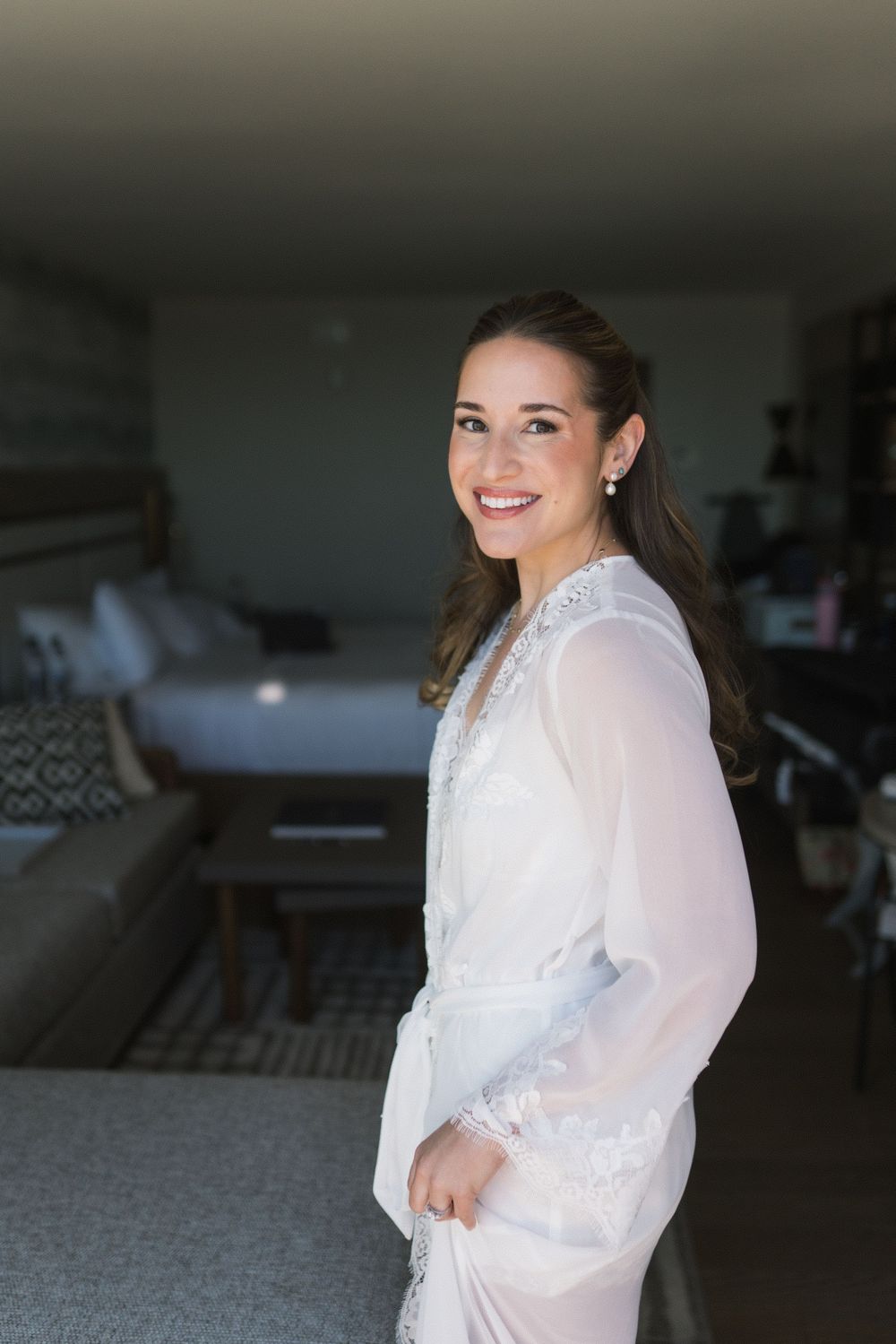 Smiling bride in elegant white lace robe posing in a hotel room on her wedding day.