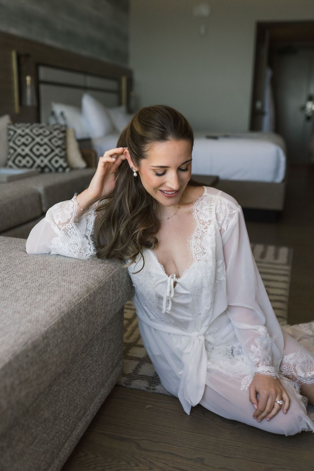Bride in white lace robe sitting on floor leaning against sofa in elegant hotel room on wedding morning.