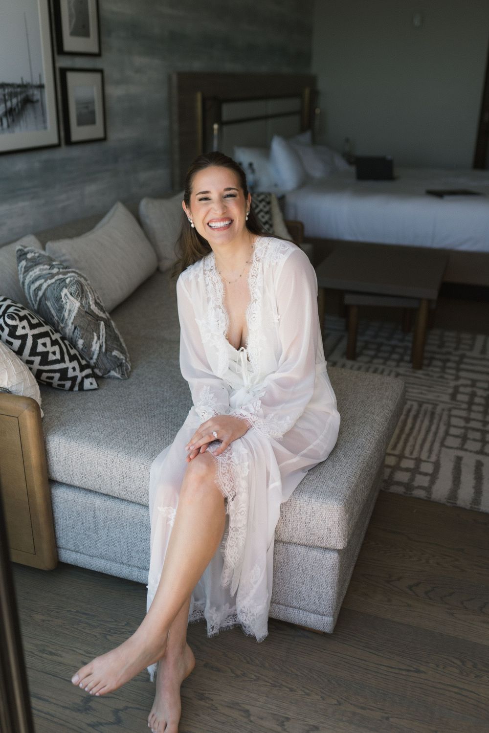 Smiling bride in white lace robe sitting on gray sofa in elegant hotel room on wedding morning.