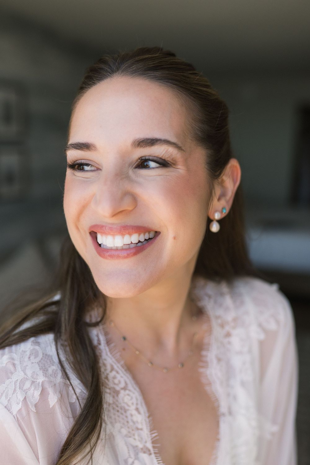 Radiant bride smiling in lace robe wearing pearl earrings and delicate necklace on her wedding day.