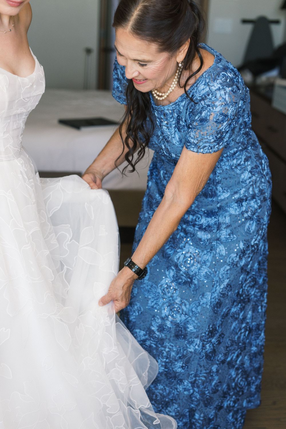 Woman in blue dress adjusting white wedding gown of bride during bridal preparations indoors.