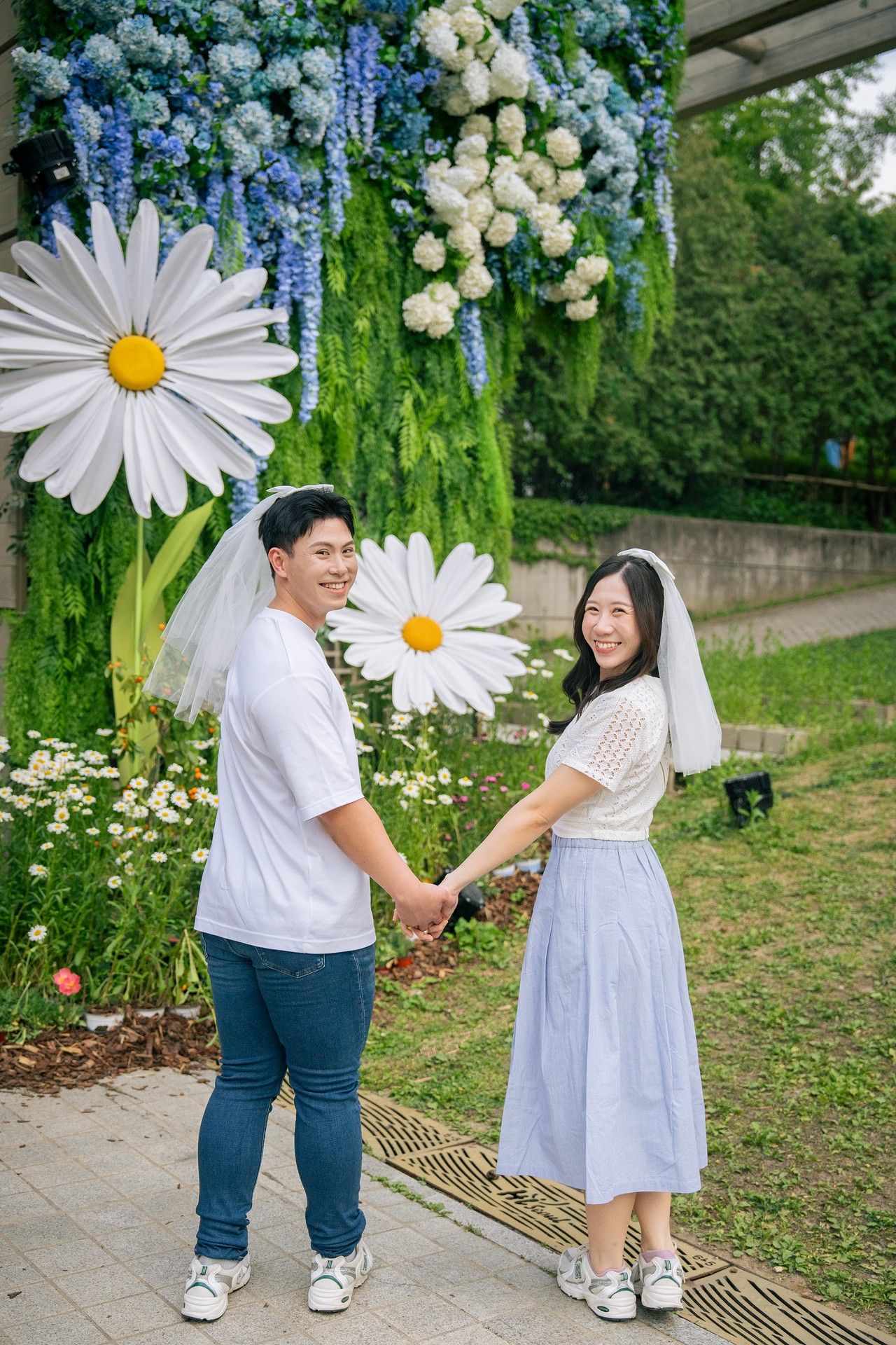 Asian couple holding hands smiling in garden with large daisy decorations and hanging blue and white flowers.