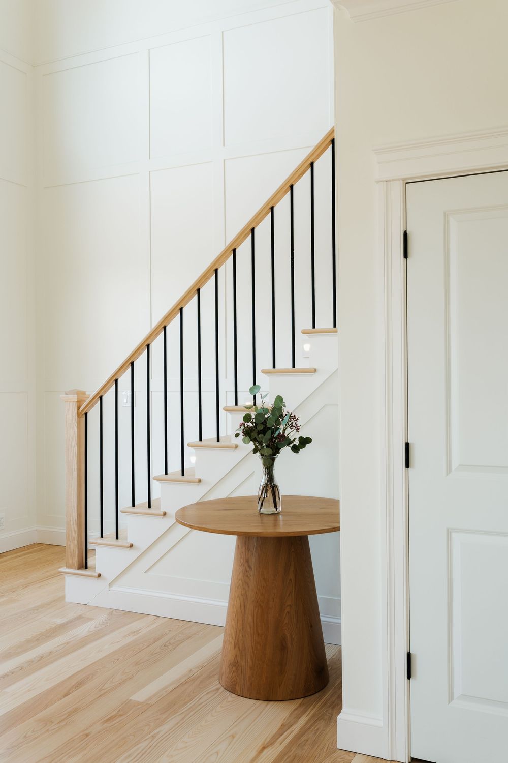 Modern foyer with wood staircase, black iron balusters, panel walls, light hardwood floors, and a round pedestal table with flowers.