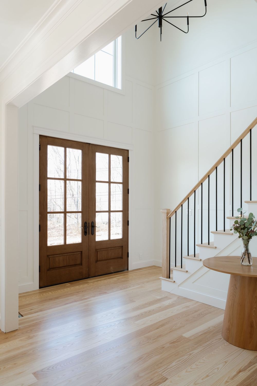 Bright foyer with wood French doors, white board and batten walls, light hardwood floors, and modern staircase with black iron balusters.