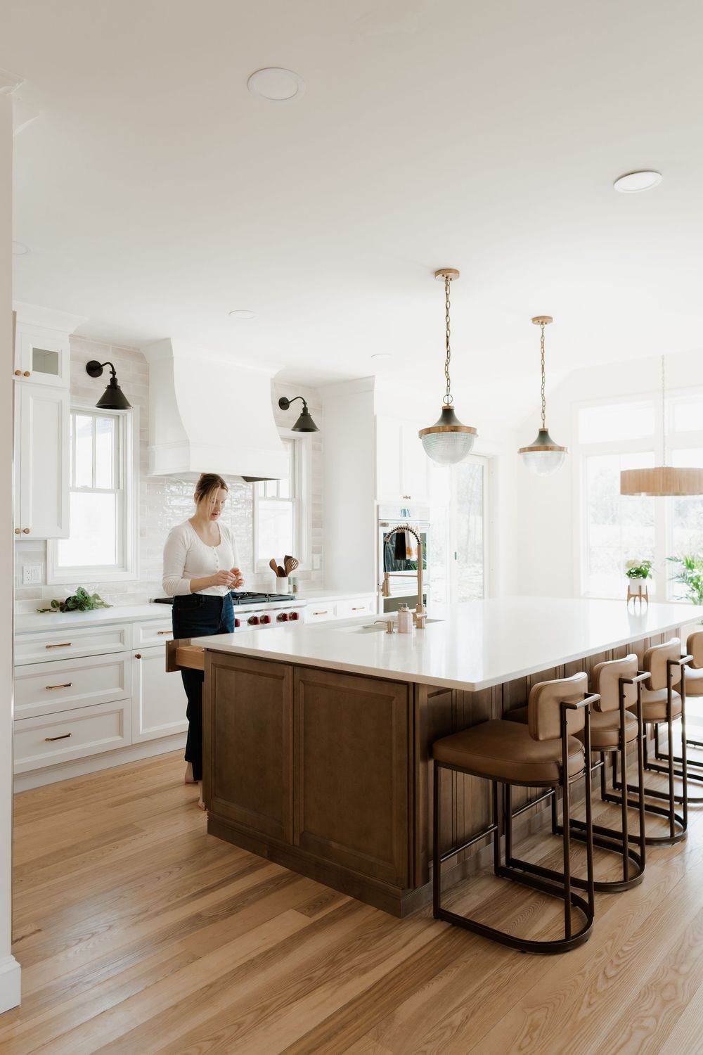 Bright white kitchen with wood island, leather barstools, pendant lights, and a woman cooking at the stove.
