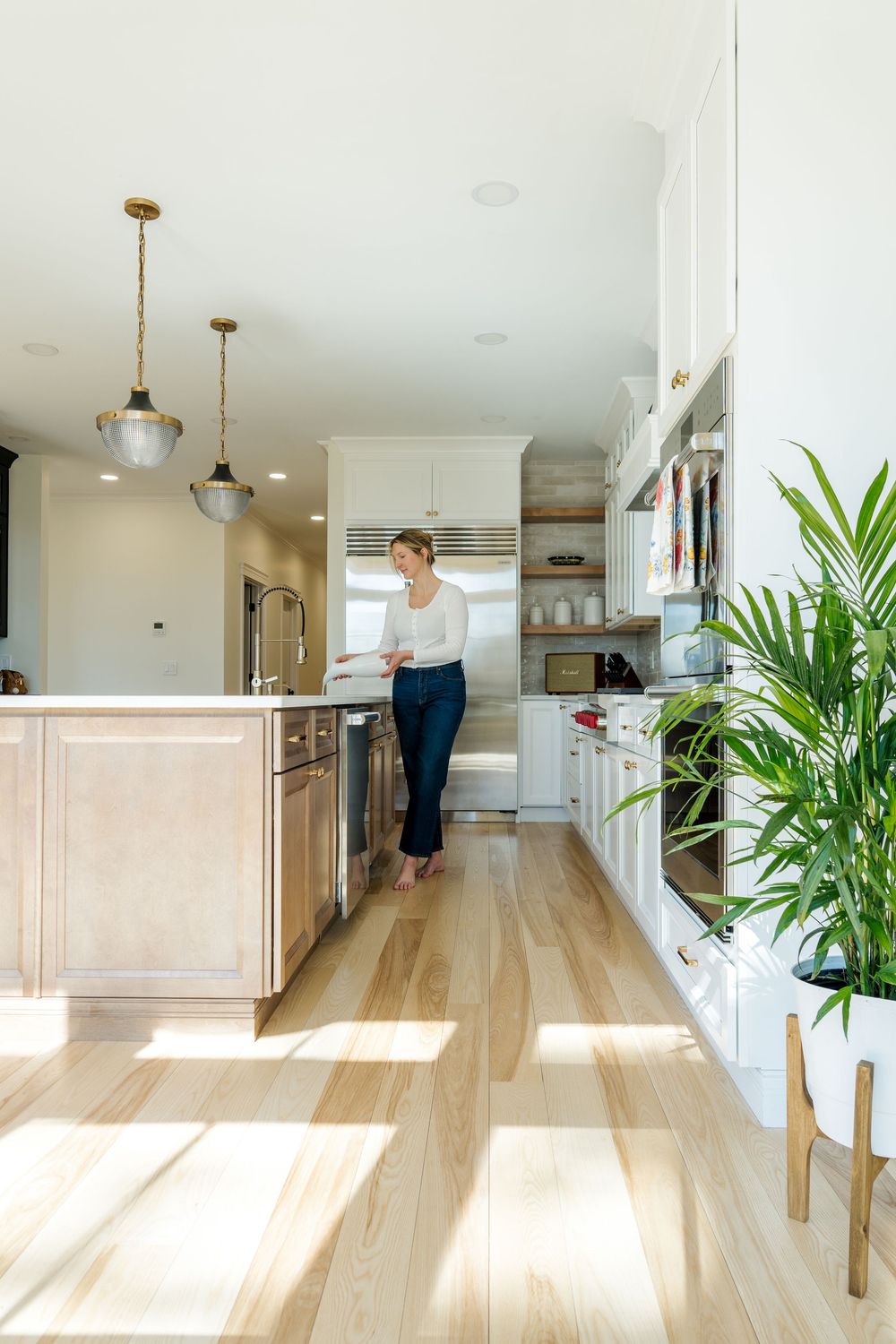 Woman standing in a bright modern kitchen with white cabinets, wood island, pendant lights, and light hardwood floors.