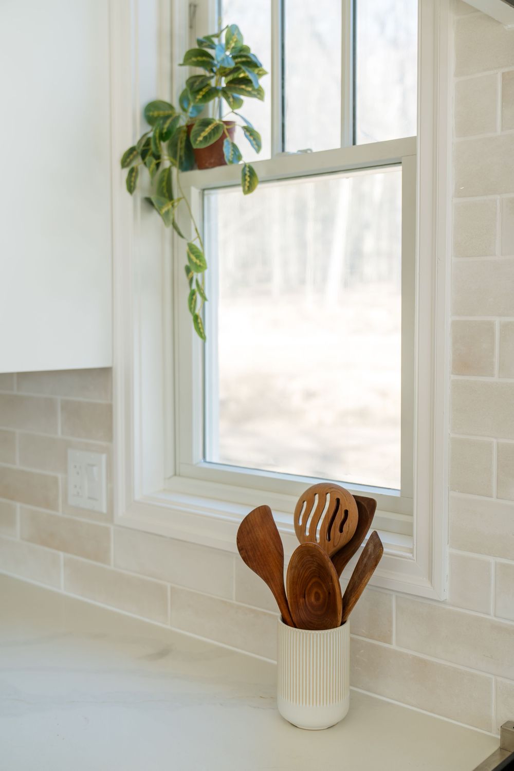 White kitchen counter with wooden utensils in a ceramic holder near a bright window with subway tile backsplash.