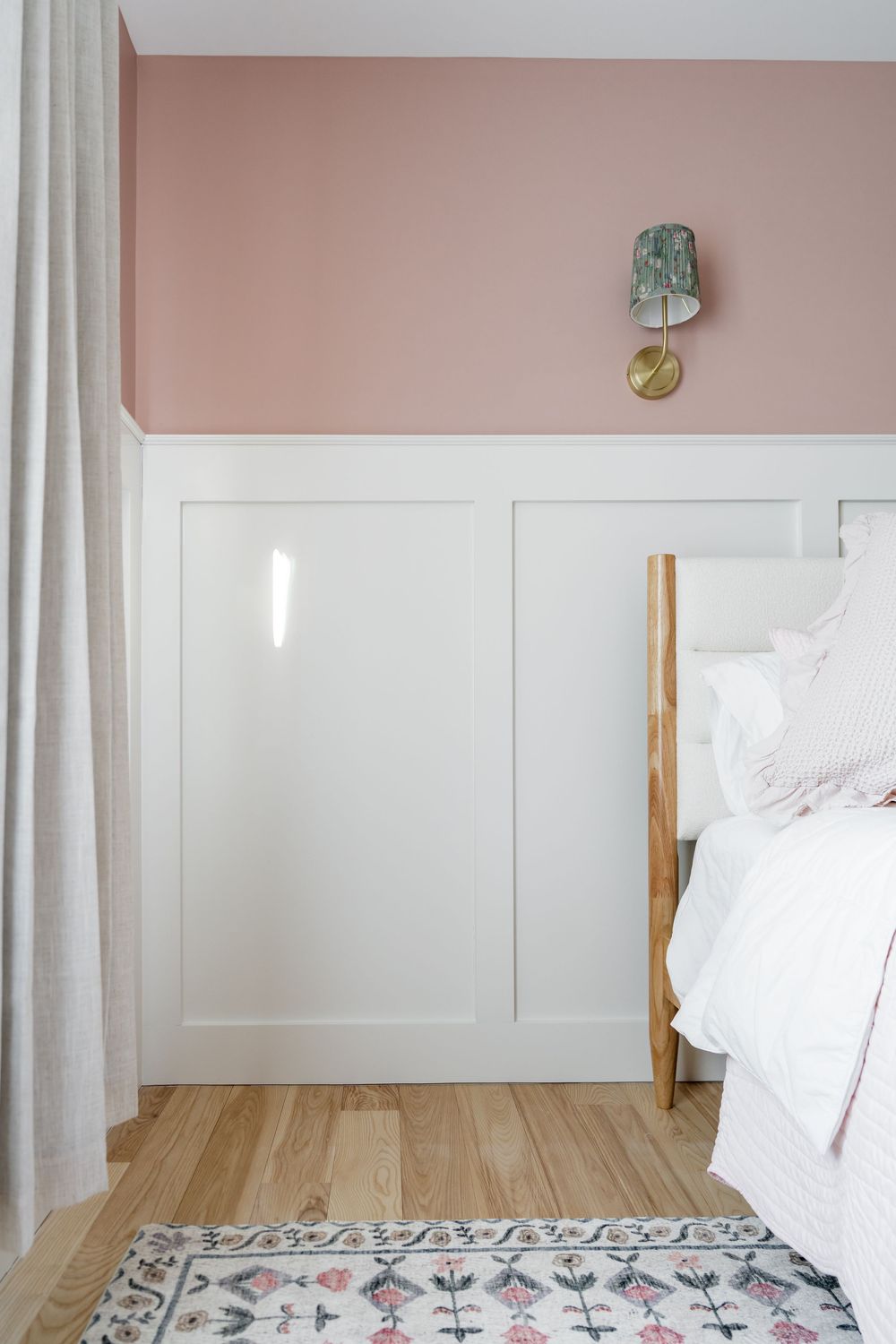 Pink and white bedroom with board and batten wainscoting, brass wall sconce, wood bed frame, and light hardwood floors.