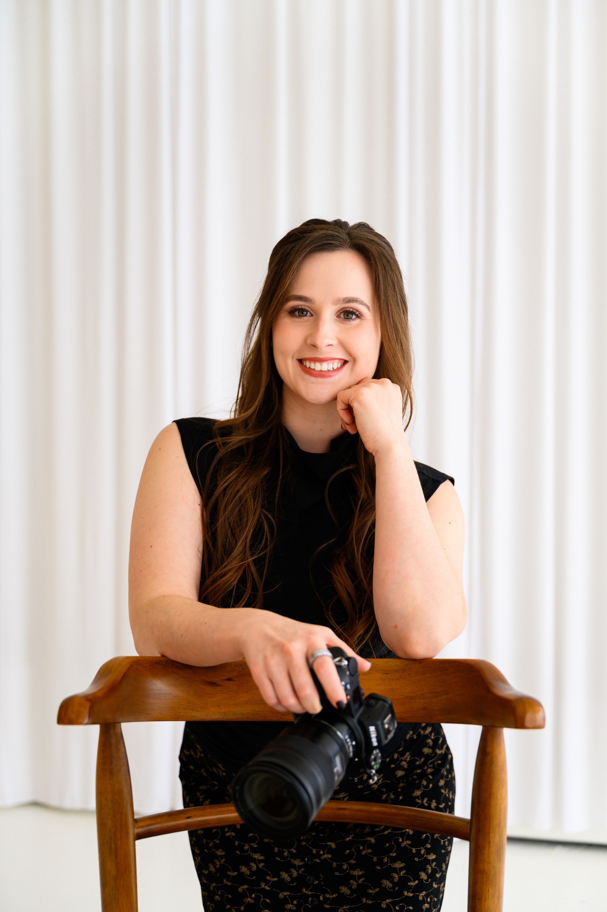 Smiling woman with long brown hair holding a Canon camera, seated on a wooden chair against a white curtain backdrop.