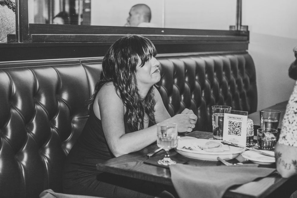 Woman sitting in leather booth at restaurant, black and white photo, dining table with glasses and plates.