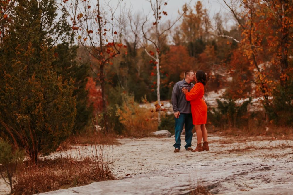 Couple kissing on a snowy path surrounded by vibrant autumn foliage in a romantic fall engagement photo.