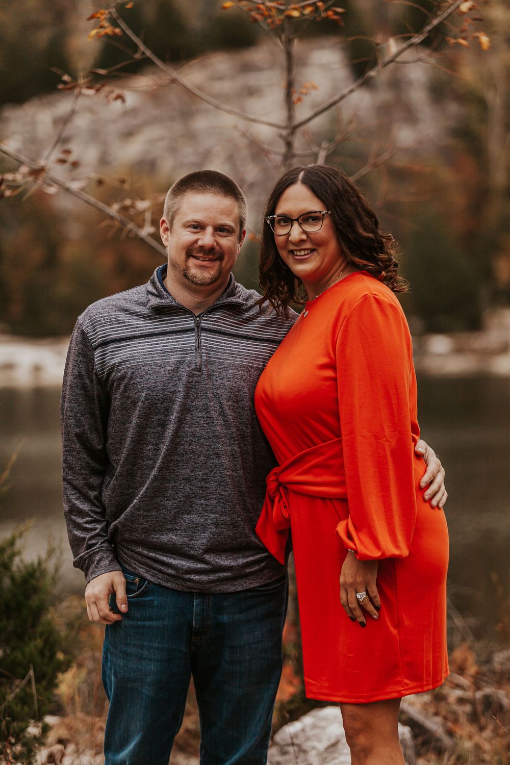Smiling couple embracing outdoors in fall setting, man in gray pullover, woman in bright orange long-sleeve wrap dress.