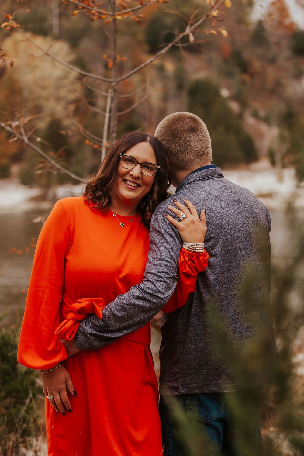Smiling woman in red dress embraces man outdoors during fall engagement photo session near snowy lake.