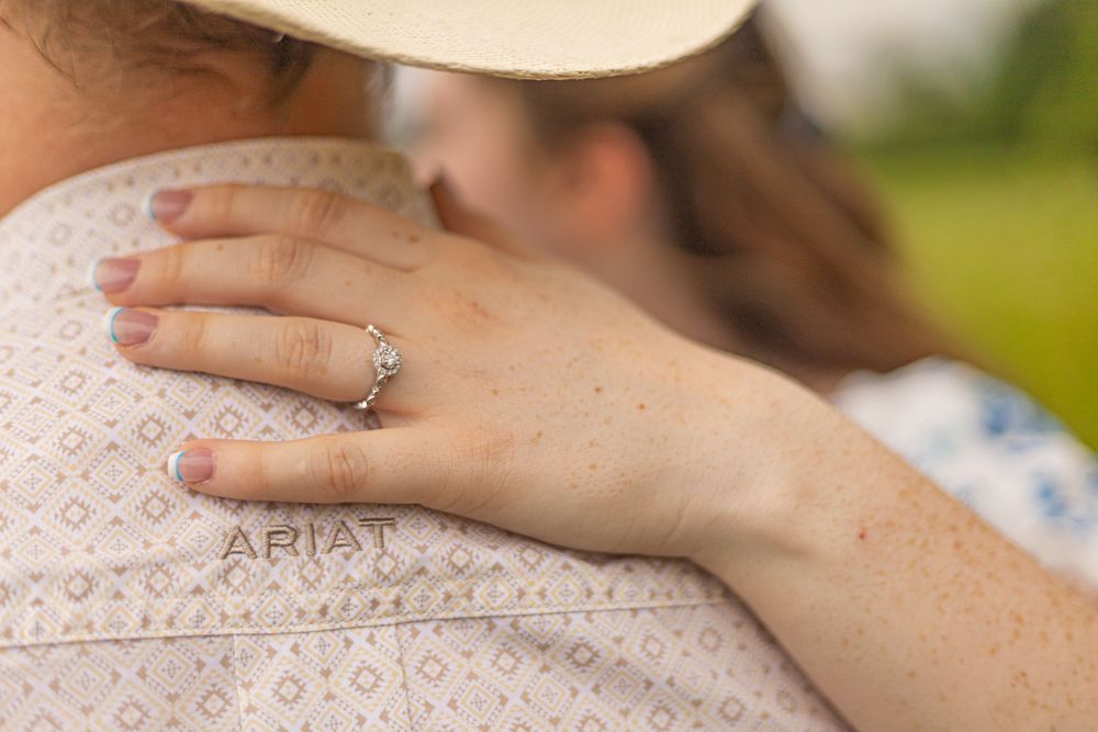 Close-up of a woman's hand with a diamond engagement ring resting on a man's Ariat shirt shoulder.