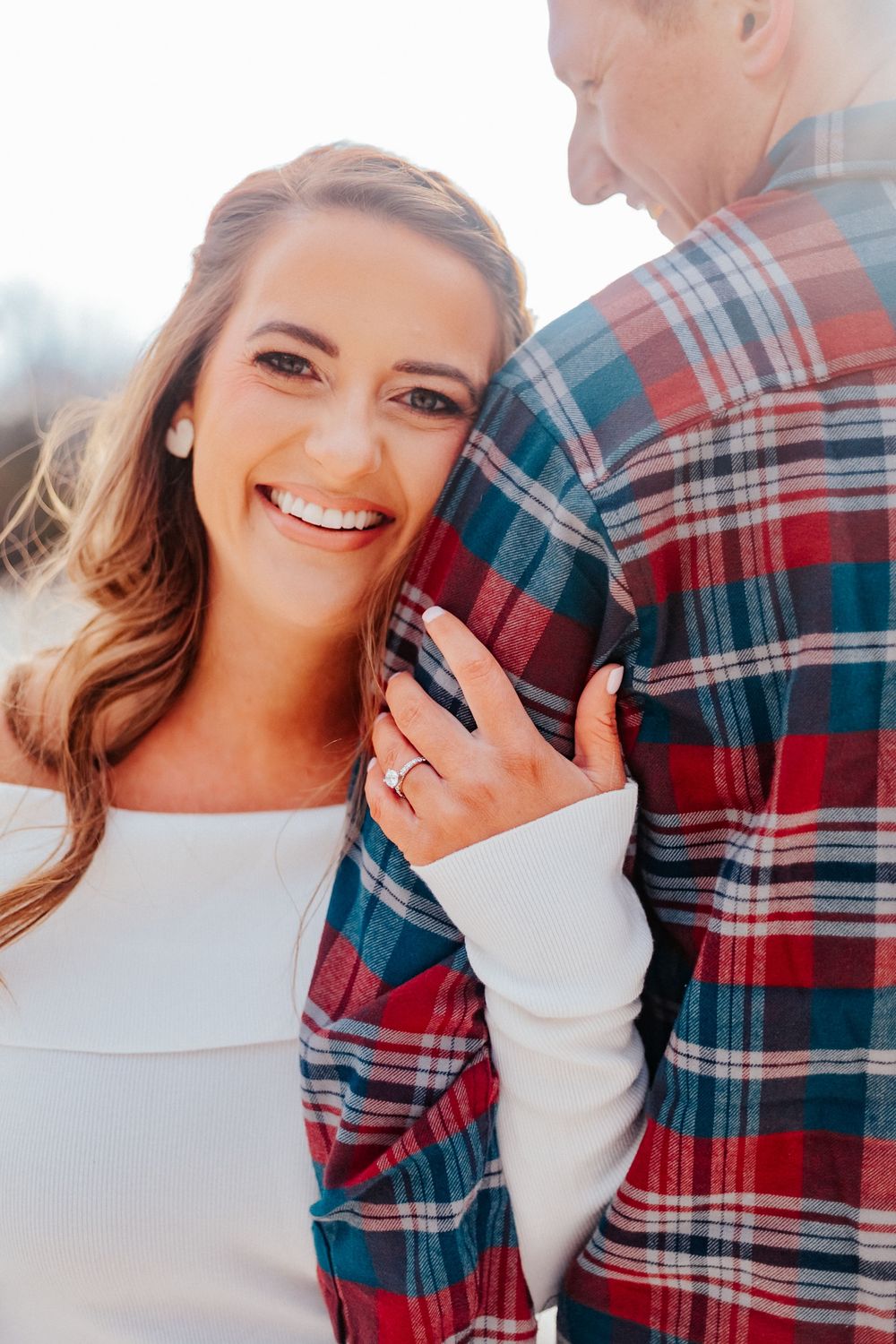 Smiling brunette woman in white top leaning against a man in red plaid shirt, showing an engagement ring.