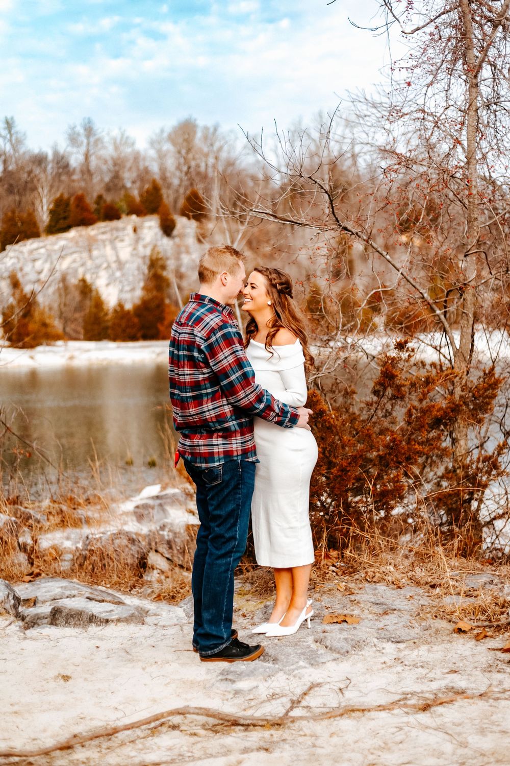 Couple embracing on rocky terrain near frozen lake surrounded by bare winter trees, woman in white dress, man in plaid shirt.