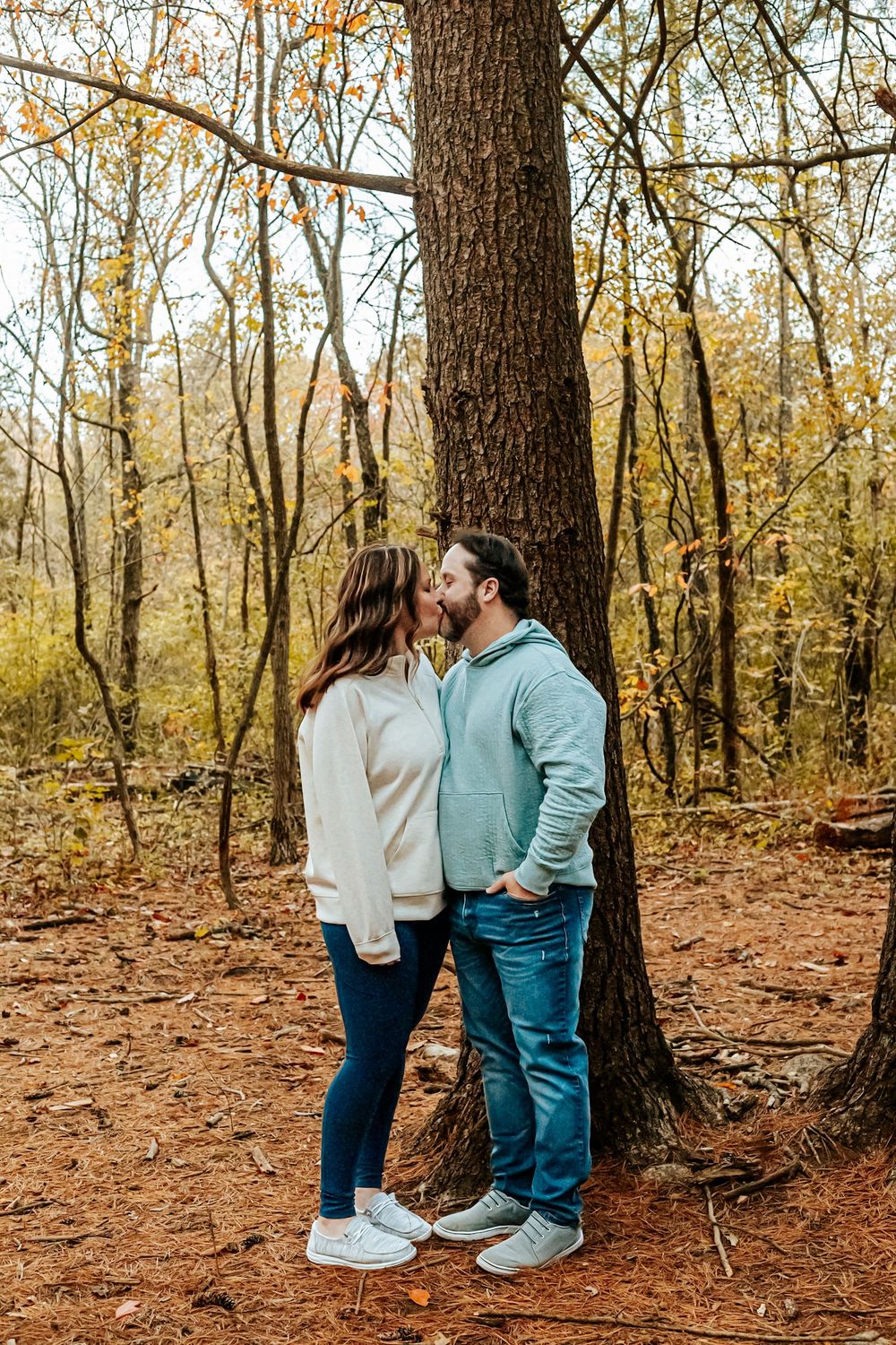 Couple sharing a kiss in an autumn forest, wearing casual sweaters and jeans, standing against a large tree.