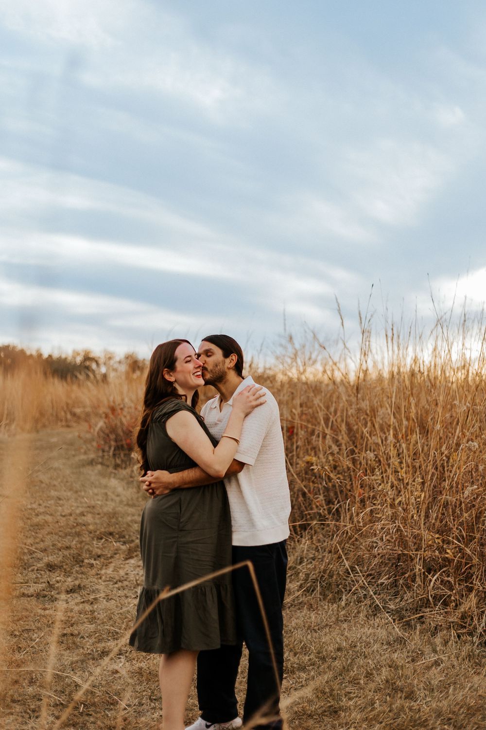 Couple embracing and kissing in a golden field, woman in olive dress smiling joyfully under a cloudy sky.
