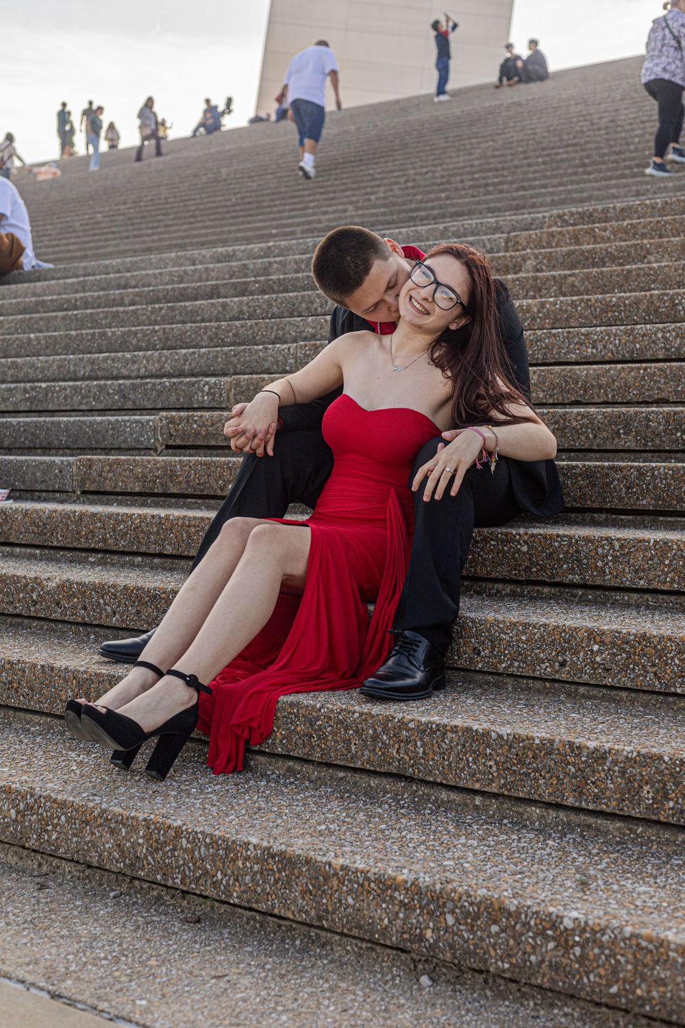 Couple in formal wear sitting on outdoor steps, woman in red dress and man in black suit embracing romantically.