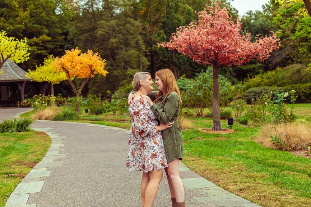 Two women embrace on a garden path surrounded by colorful autumn trees in a scenic park setting.