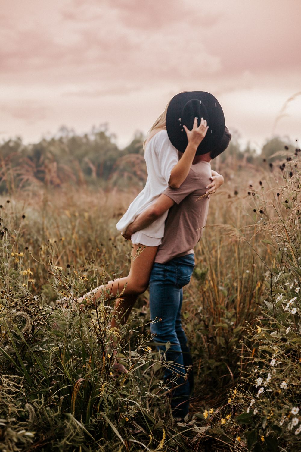 Couple embracing romantically in a wildflower field, woman in hat lifted by man in jeans under a pink cloudy sky.