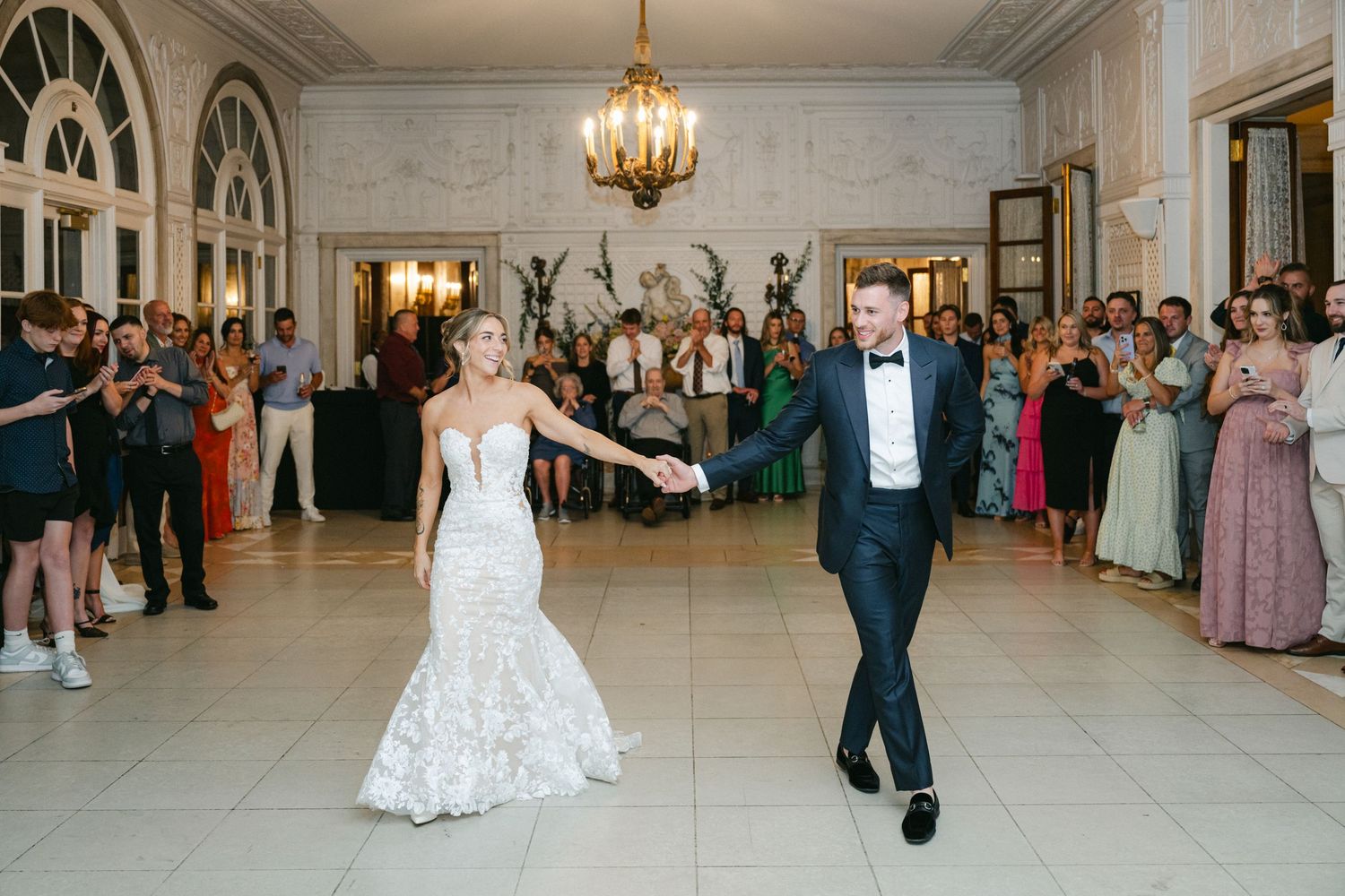 Bride in mermaid gown and groom in tuxedo share first dance at elegant ballroom wedding reception.