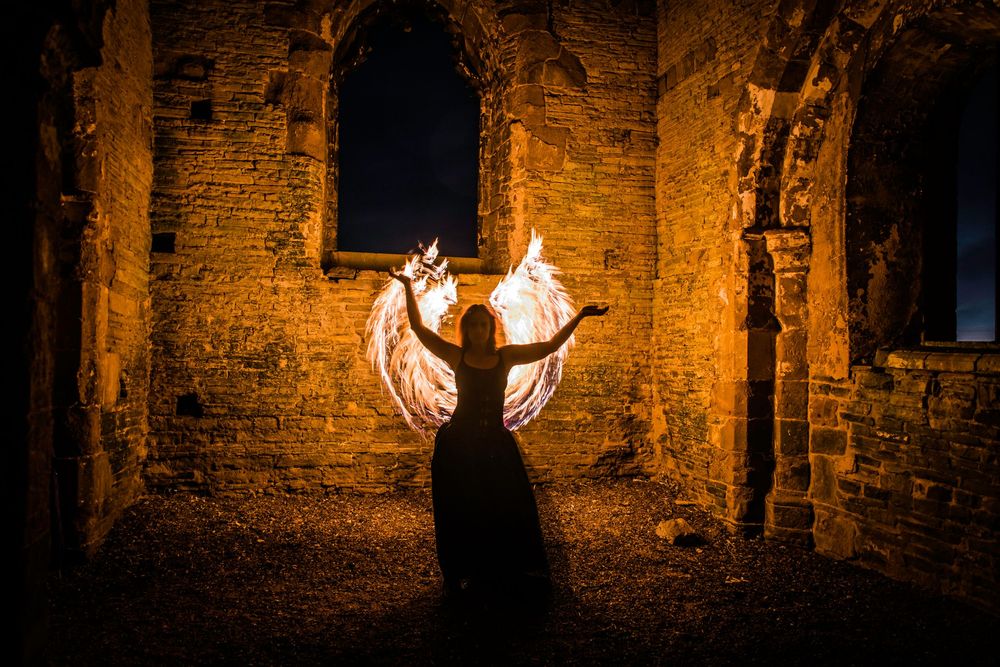 Person holding fire wings in ancient ruins at night, creating dramatic orange glow against stone walls.