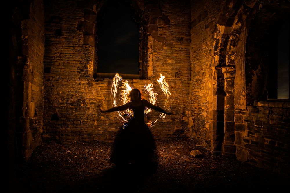 Silhouetted fire dancer performing with flaming wings in ancient ruins at night.