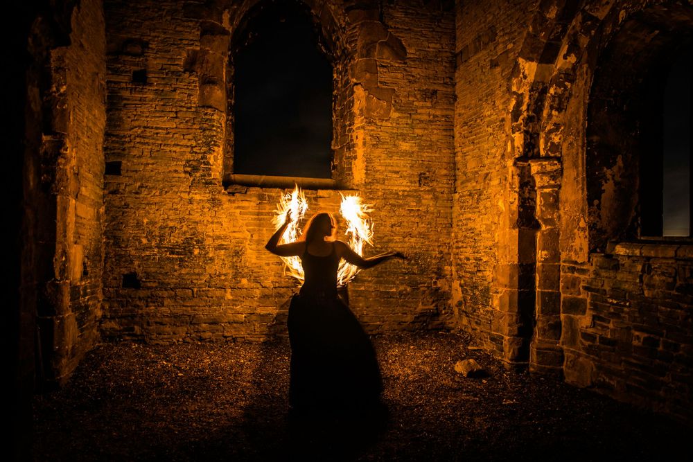 A mysterious woman in a black gown holds fire in ancient stone ruins, bathed in dramatic orange light.