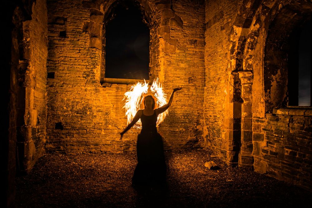 Woman in black dress performing fire dance in ancient stone ruins at night with dramatic orange flames.