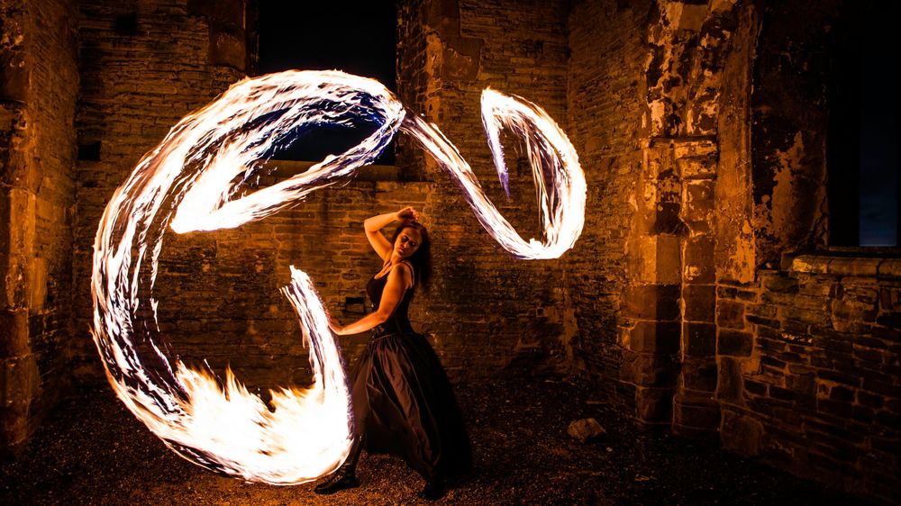 Woman performing fire dancing in ruins at night, creating glowing spiral light trails with flames in a long exposure photograph.