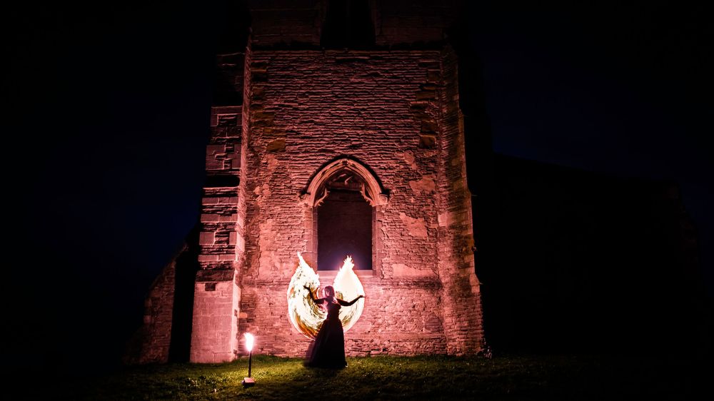 Fire performer twirling glowing wings at night before an ancient stone church tower illuminated in red light.