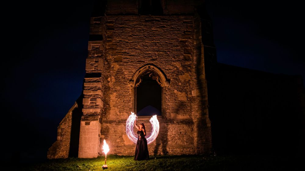 Fire performer creating glowing circles at night before an ancient stone church illuminated by torchlight.