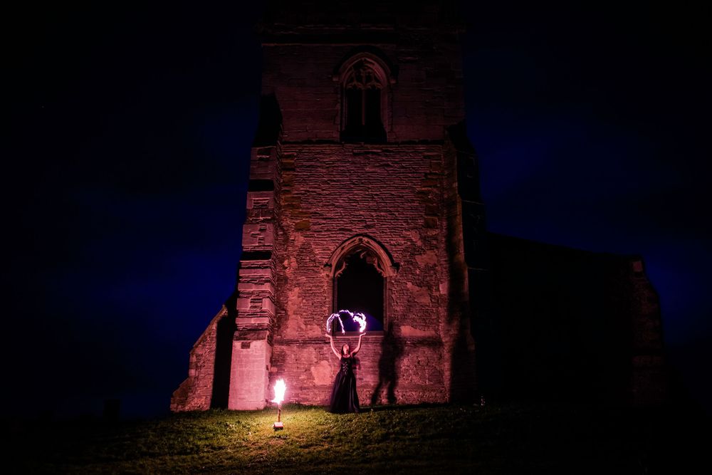 Fire performer with glowing orb beside illuminated medieval stone church tower at night.
