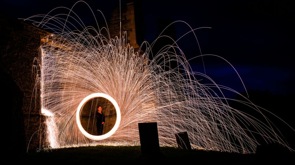 Steel wool spinning creates a circle of golden sparks against a dark night sky near an old stone building.