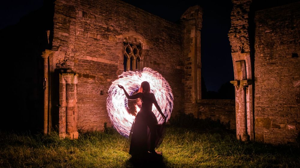 Woman spinning fire poi creating a glowing circle of light in front of an ancient stone building at night.