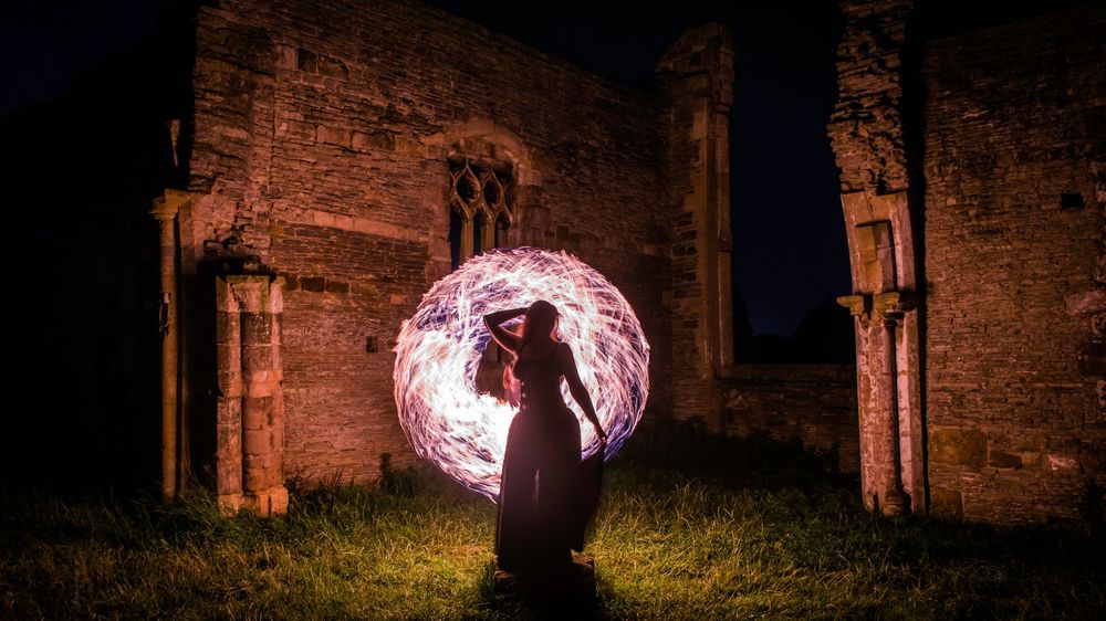 Silhouette of a person spinning a glowing orb of light in front of ancient stone ruins at night.