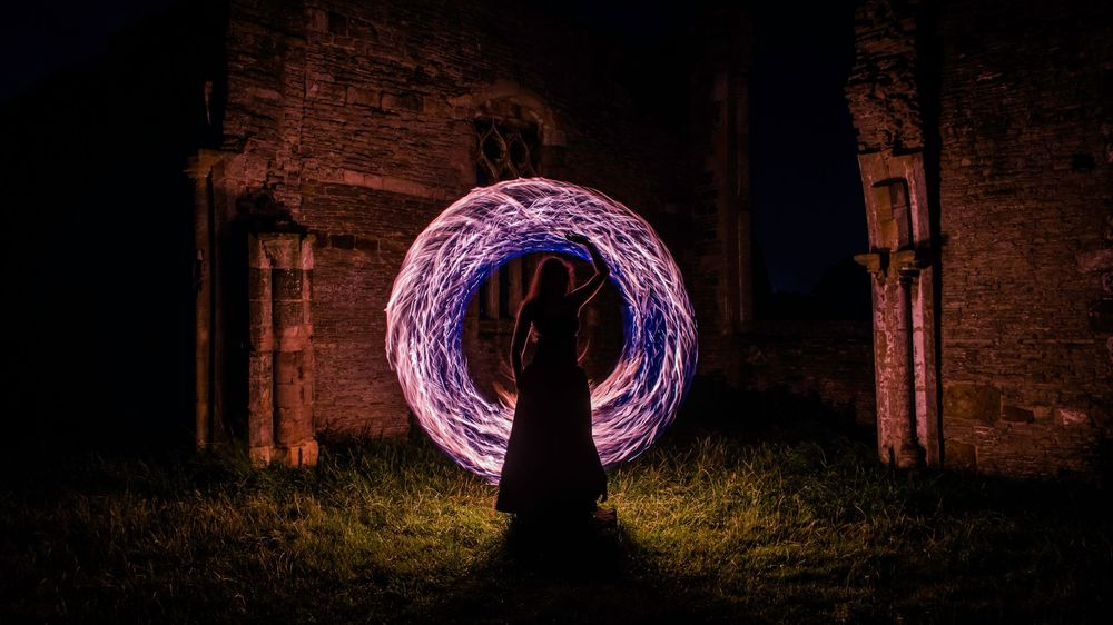 Person creating glowing purple light orb at night near ancient stone ruins, long exposure photography art.