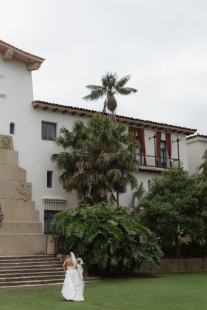 Bride in white wedding gown and veil walks across green lawn near white Spanish-style building with palm trees.