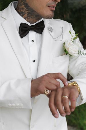 Tattooed groom in white tuxedo with black bow tie adjusting gold cufflinks, wearing rings and white floral boutonniere.