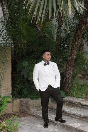 Groom in white tuxedo jacket and black bow tie poses on stone steps surrounded by tropical palm trees.