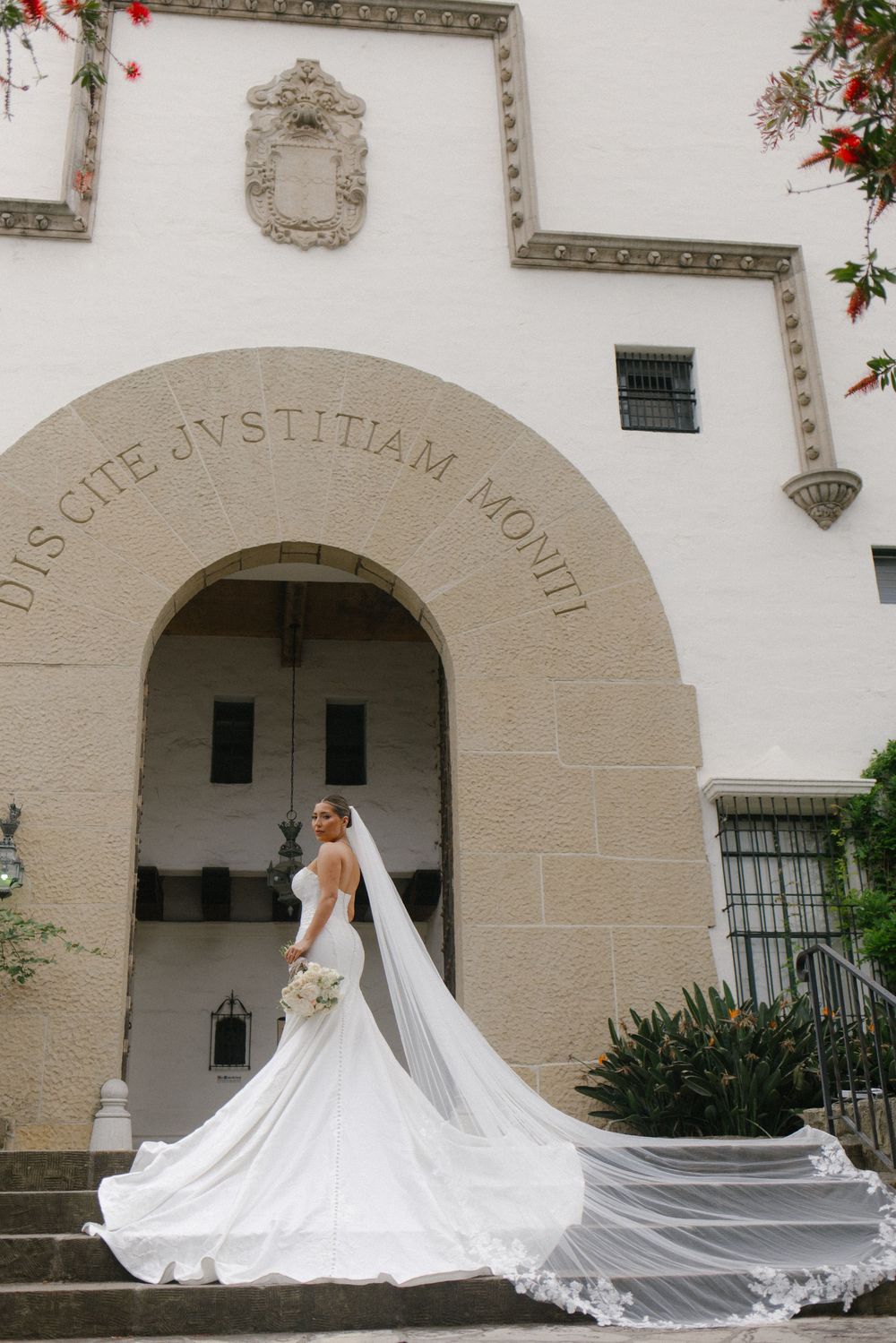 Bride in elegant white gown with cathedral veil poses on steps before arched entrance of historic building.