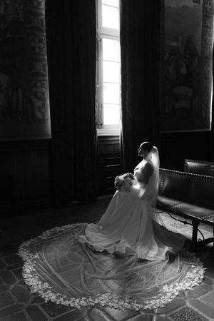 Black and white photo of a bride in a flowing gown with lace veil train, standing by a window in an elegant room.