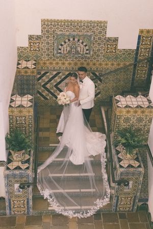 Bride and groom embrace on ornate tiled staircase, bride wearing cathedral veil and holding white bouquet.